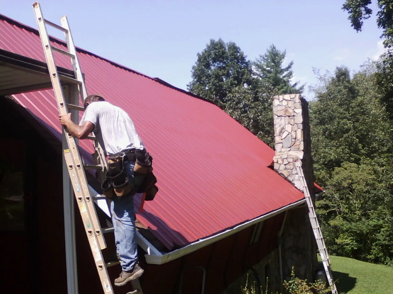 Roofer on a ladder beside a red metal roof and stone chimney — Soffit & Fascia Repair by Roofing Brothers in Haworth